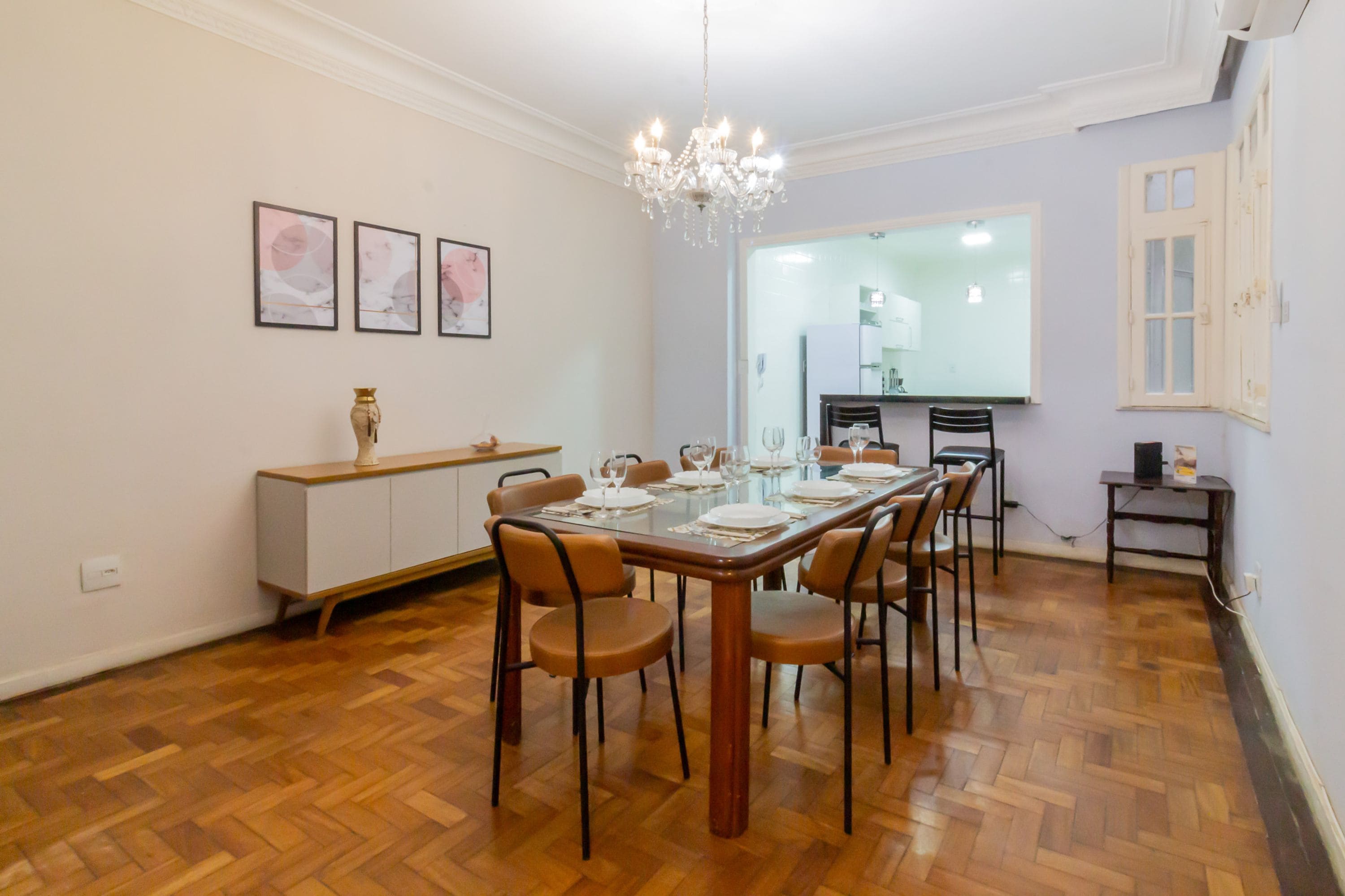 A dining area with a wooden table set for six, surrounded by chairs. There is a chandelier above the table, and a white cabinet with a vase on top is visible to the left.