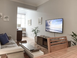 Uma sala de estar moderna com televisão de tela plana na parede, um sofá, uma mesa de centro e um espaço de jantar com uma cadeira de madeira.