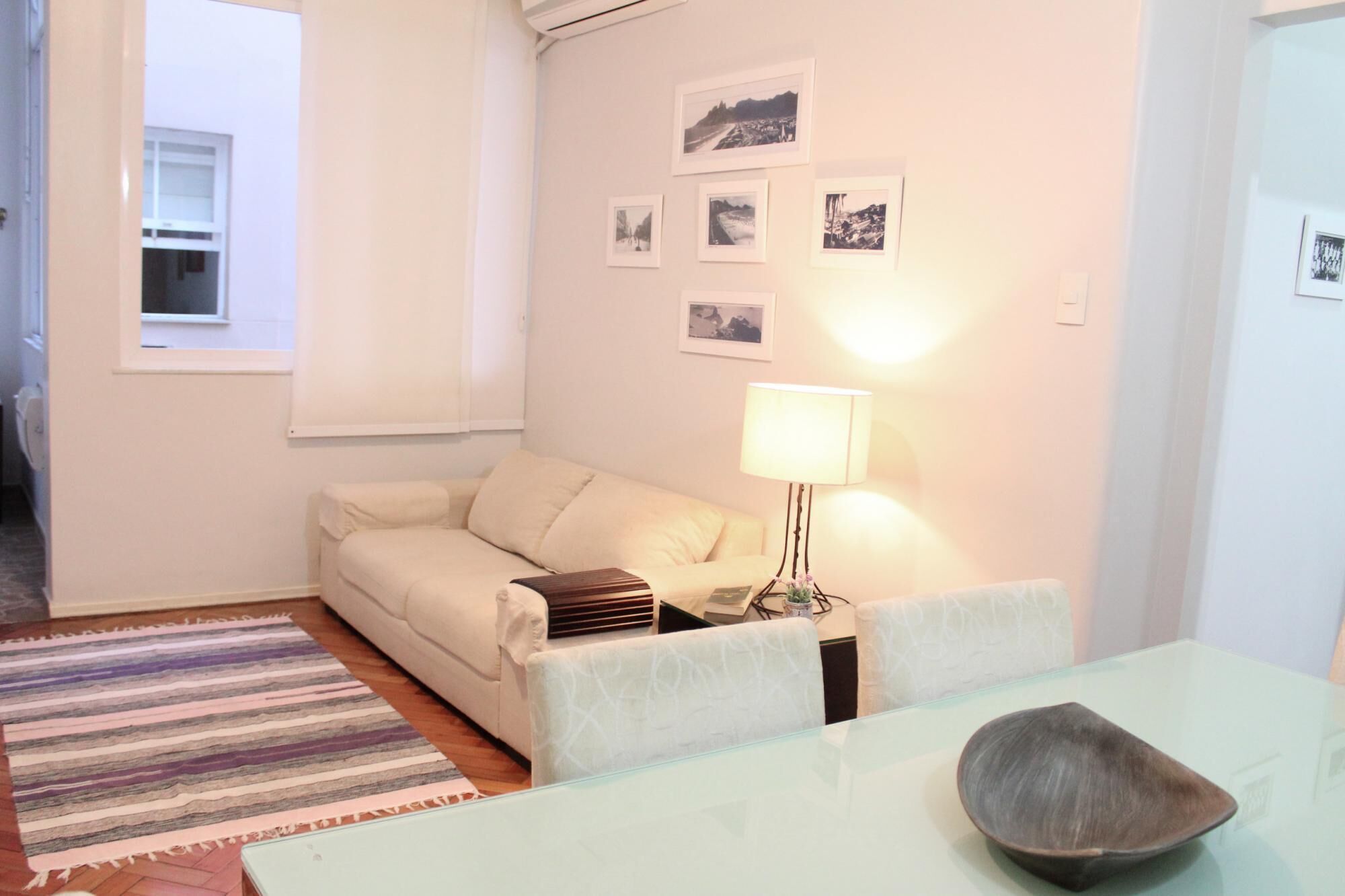 A living room with a sofa, a glass table, a lamp, and a bowl. There are pictures on the wall and a window with blinds.