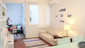 Un salon moderne avec un canapé, une table basse en verre et une lampe. Un tapis à rayures recouvre le sol, et une fenêtre est équipée de stores.