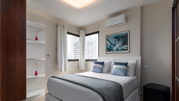A bedroom with a bed, white walls, a painting, a window, and a built-in shelf with a vase. - Attractive in Barra da Tijuca With a View As1602 (Rio de Janeiro)