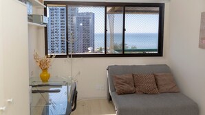A small, modern room with a grey sofa, a glass table with a yellow vase, and a window overlooking a building and the sea. - Attractive in Barra da Tijuca With a View As1602 (Rio de Janeiro)