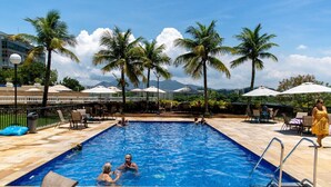 A swimming pool with palm trees, lounge chairs, and umbrellas. - Attractive in Barra da Tijuca With a View As1602 (Rio de Janeiro)
