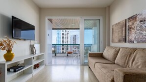 A modern living room with a flat-screen TV, a sofa, and a balcony with a view of high-rise buildings. - Attractive in Barra da Tijuca With a View As1602 (Rio de Janeiro)
