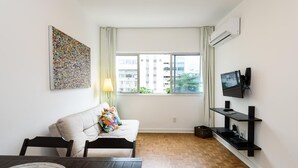 Ein modernes Wohnzimmer mit einer Couch, einem Esstisch, einem an der Wand befestigten Fernseher und einem Fenster mit Blick auf Gebäude.