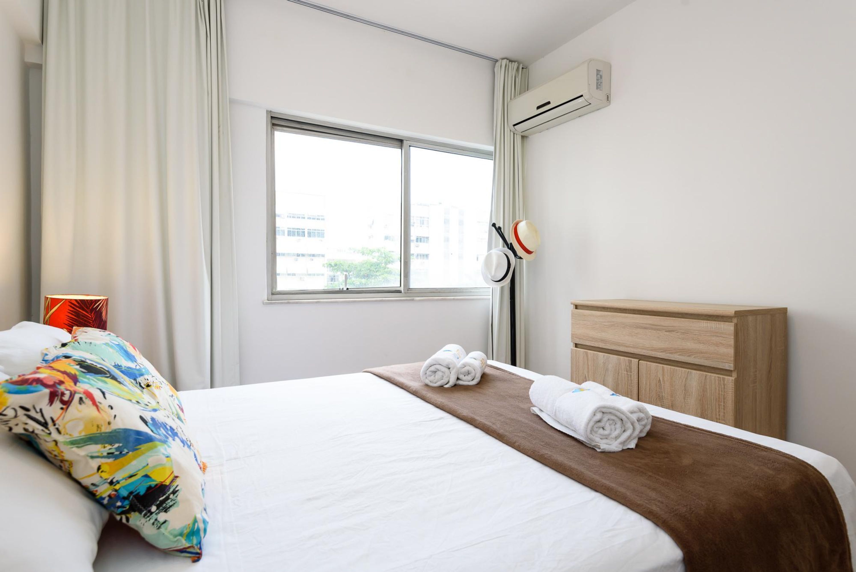A hotel room with a large bed, a window with curtains, a wooden headboard, and a wall-mounted air conditioner.