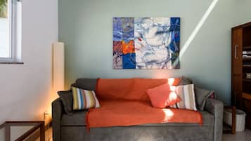A living room with a grey sofa, orange throw blanket, and a colorful abstract painting on the wall.