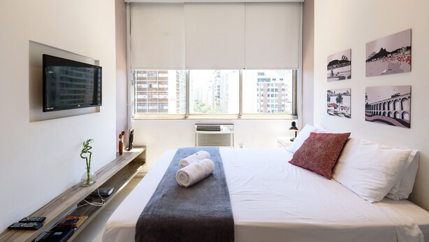 A modern hotel room with a large bed, a flat-screen TV mounted on the wall, a window with a city view, and artwork on the wall.