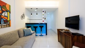 A modern living room with a sofa, a TV, a wooden cabinet, and a kitchen area with stools and a bicycle.