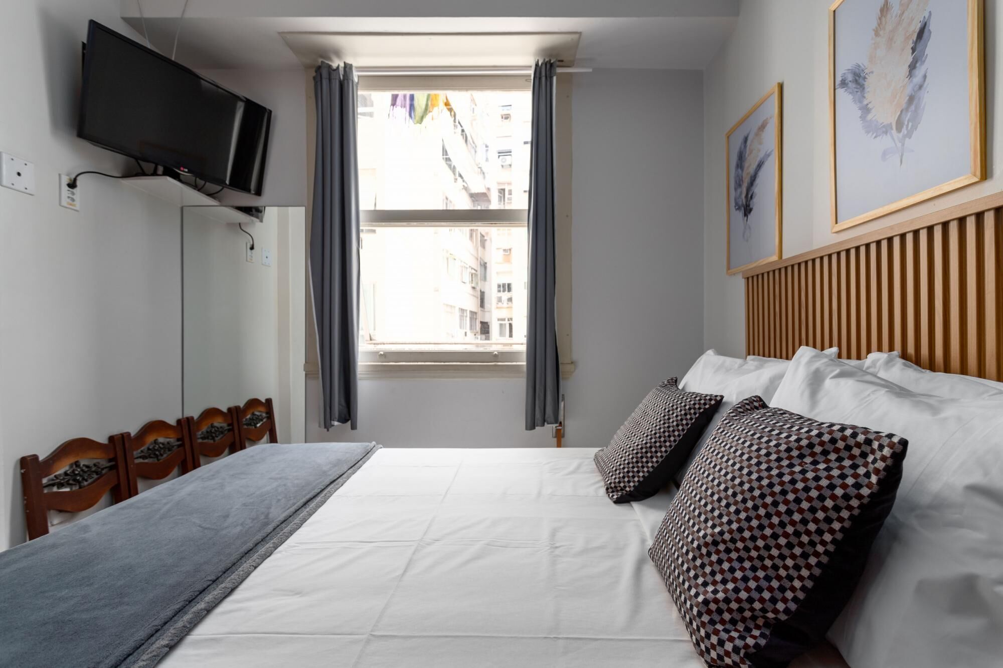 Une chambre à coucher avec un lit, un téléviseur fixé au mur, une fenêtre avec des rideaux et une vue sur les bâtiments à l’extérieur.
