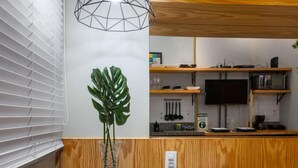 Una stanza compatta con una parete in legno decorativa, un tavolino con una pianta, un letto con biancheria bianca e un angolo cottura con TV e microonde.