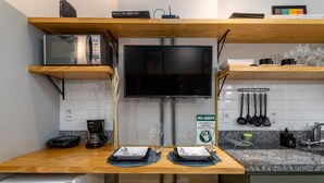 Una cucina compatta con forno a microonde, tostapane e macchina per il caffè sul piano di lavoro, una TV a schermo piatto montata sopra un tavolo da pranzo con sgabelli gialli, e un cartello che promuove pratiche ecologiche.