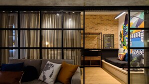 A modern living room with a grey sofa, a coffee table, and a sliding glass door leading to a dining area with a brick wall and a colorful mural. - 5 Minutes From Copacabana Beach Nsc902 (Rio de Janeiro)