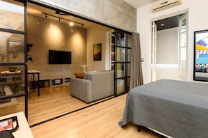A modern hotel room with a bed, a sofa, a TV, and a glass door leading to a balcony.