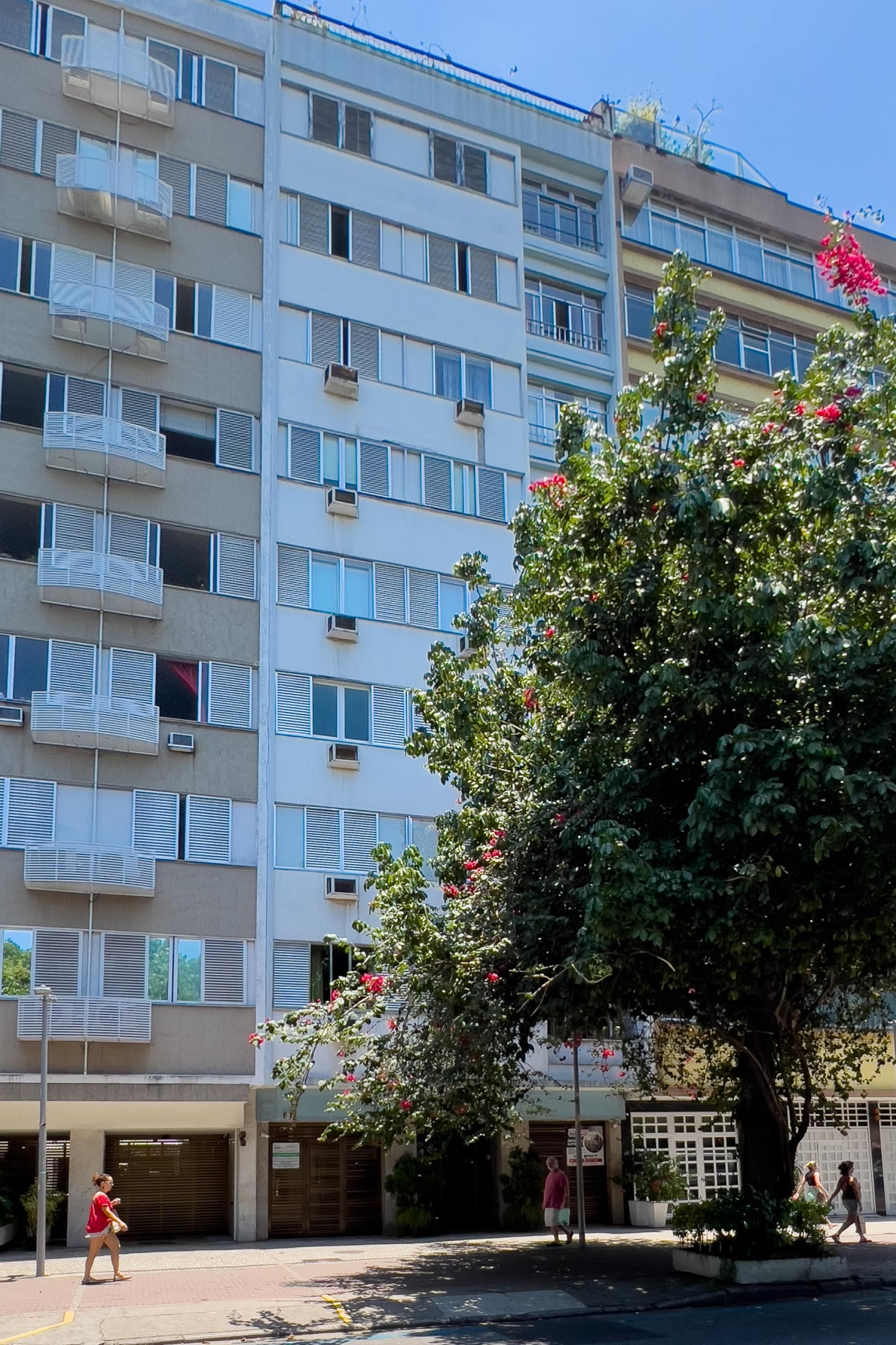 A multi-story residential building with balconies, a tree with pink flowers, and a pedestrian walkway.