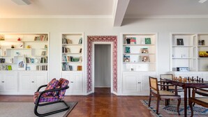 Una habitación amplia con piso de madera, paredes blancas y una gran estantería llena de libros y objetos decorativos. Un comedor con sillas se encuentra cerca de la estantería.