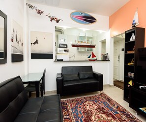 Ein modernes Wohnzimmer mit einer schwarzen Couch, einem Glastisch, einem gemusterten Teppich, einer Küchenzeile mit einer Mikrowelle und einem Bücherregal mit verschiedenen Gegenständen.