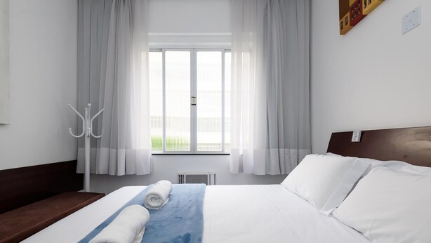 Quarto de hotel com cama grande, roupa de cama branca, almofada azul de destaque e vista para área externa verde através de uma janela com cortinas brancas.