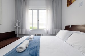 Ein Hotelzimmer mit einem großen Bett, weißen Bettwäsche, einem blauen Zierkissen und Blick auf eine grüne Außenanlage durch ein Fenster mit weißen Vorhängen.