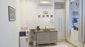 Cozinha compacta com micro-ondas integrado, máquina de café e um pequeno frigorífico. Há uma janela com cortinas e vista para o exterior.