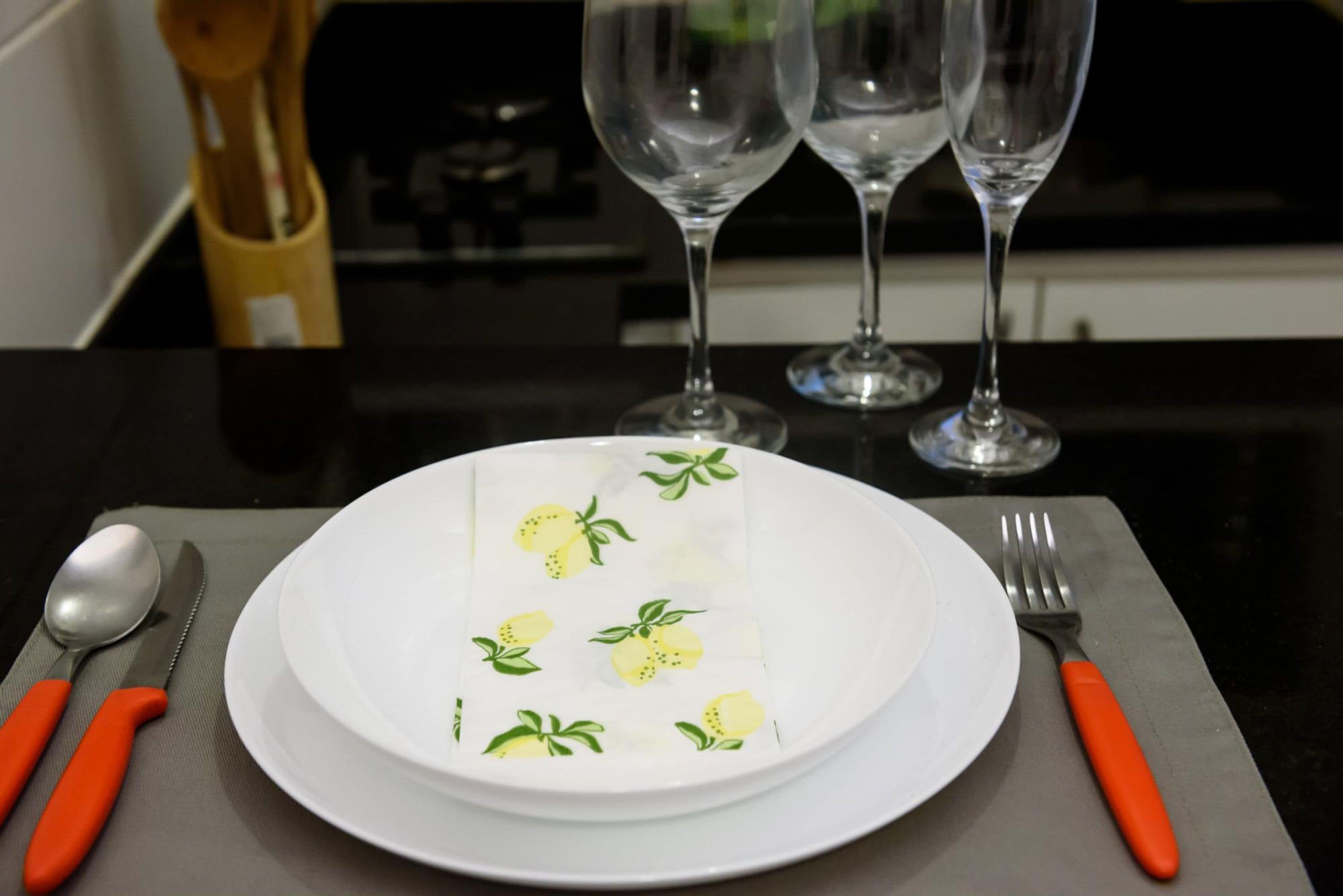 Une table à manger avec une assiette blanche ornée de motifs citronnés, accompagnée d’une cuillère, d’un couteau, d’une fourchette et de deux verres à vin vides.