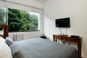 A bedroom with a large bed, a TV on the wall, and a window with a view of greenery. - Peace in Copacabana Forest View Br1009 (Rio de Janeiro)