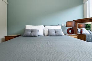 A neatly made bed with a grey quilt, two white pillows, and two grey pillows. There is a wooden nightstand with a lamp and a mirror. The wall is painted in a light teal color. - Peace in Copacabana Forest View Br1009 (Rio de Janeiro)