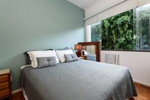 A bedroom with a bed, a nightstand, a mirror, and a window with a view of greenery. - Peace in Copacabana Forest View Br1009 (Rio de Janeiro)