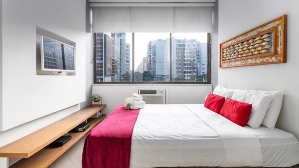 Ein modernes Hotelzimmer mit einem Bett, einer Holzbank, einem Fenster mit Blick auf die Stadt und einem gerahmten Kunstwerk an der Wand.