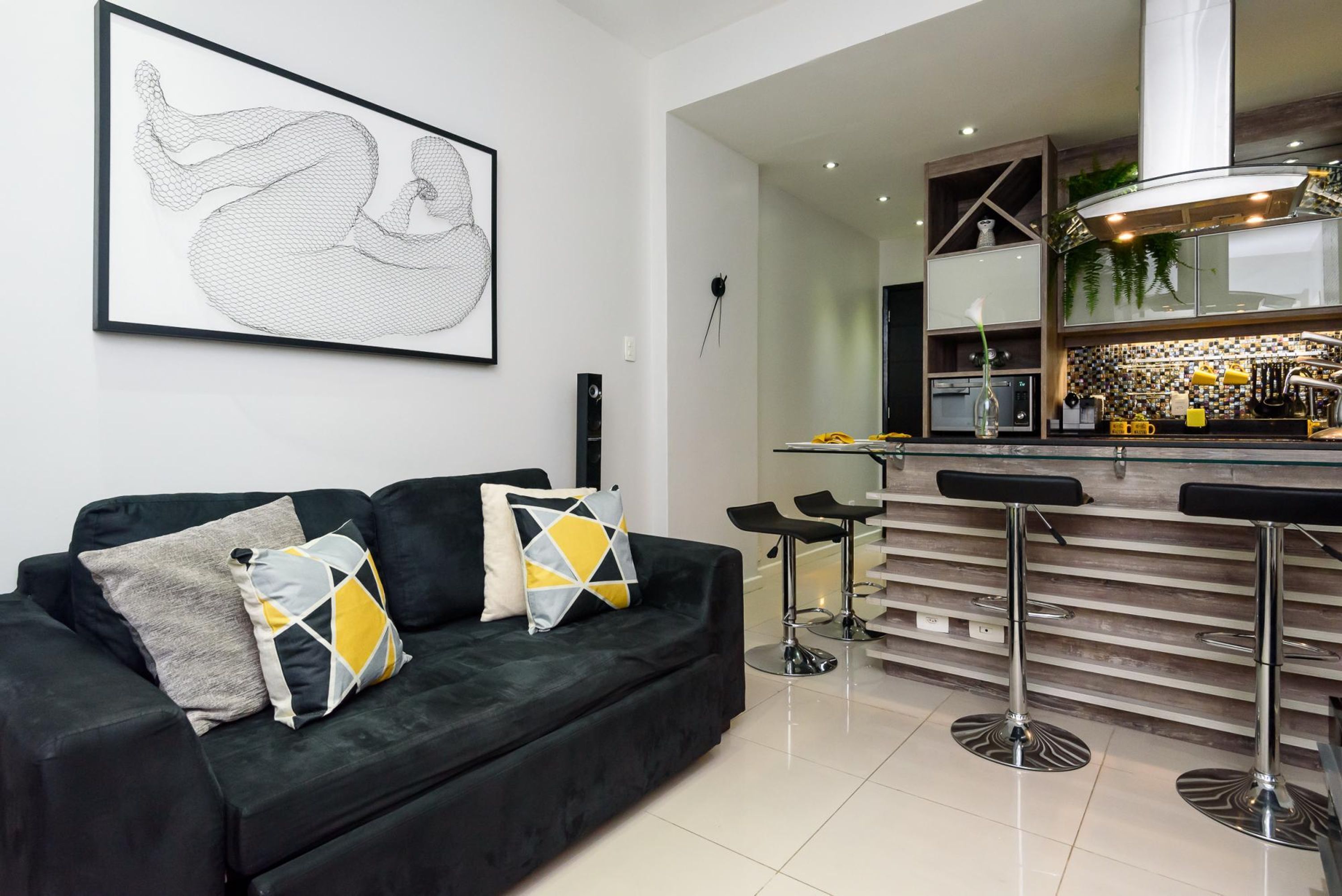 A modern living room with a black sofa, two chairs, a glass coffee table, and a kitchen area with a bar counter and stools.