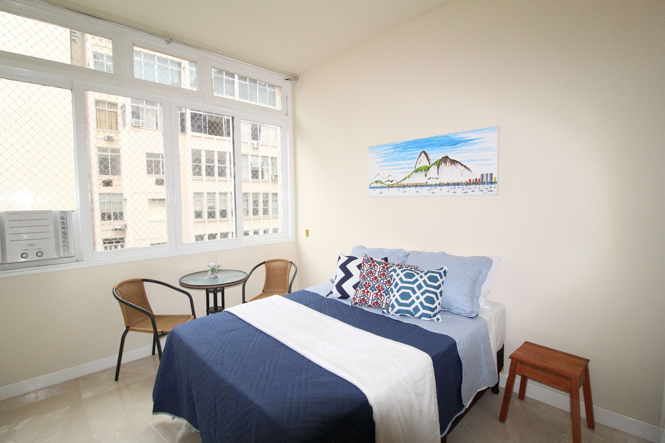 A bedroom with a bed, a chair, a small table, and a window with a view of buildings.