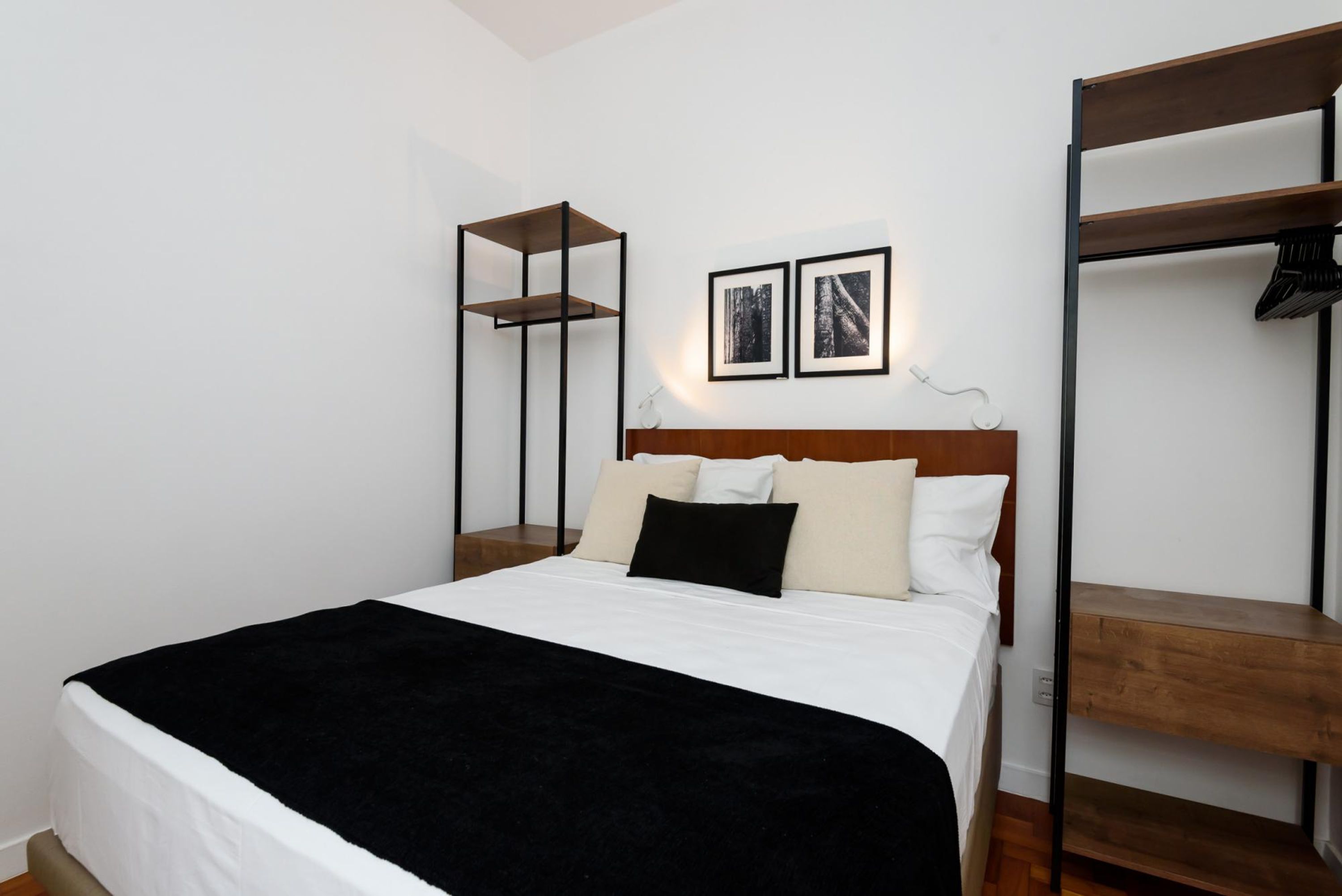 A bedroom with a bed, a bedside table, a wall-mounted shelf, and two framed pictures.