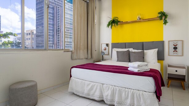 Quarto moderno com uma janela ampla, uma cama com cobertor vermelho, uma mesa de cabeceira e uma pequena planta em uma prateleira.