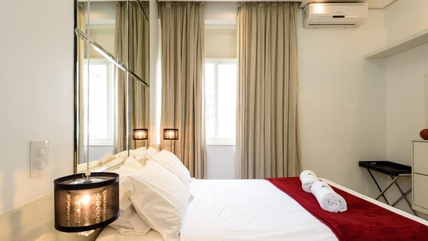 A hotel room with a bed, a red blanket, a mirror, a wall-mounted air conditioner, and a window with curtains. - Style in Ipanema Good Location Vp305 (Rio de Janeiro)