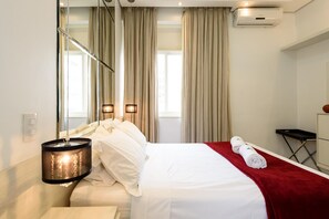 Habitación de hotel con cama, manta roja, espejo, aire acondicionado de pared y ventana con cortinas.
