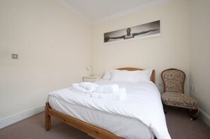 A neatly made bed with white linens and towels, a nightstand with a lamp, a floral armchair, and a painting on the wall.