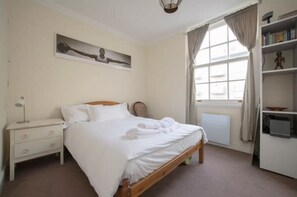 A bedroom with a bed, a nightstand, a lamp, a window with curtains, and a bookshelf. - Charming 1BD Victorian Terrace Flat in Dalston (London)
