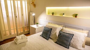 A neatly arranged bedroom with a bed, pillows, a bedside table with a lamp, a small plant, and a wall-mounted shelf with additional plants.