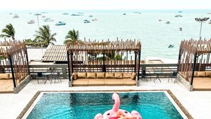 A swimming pool with a pink flamingo float, surrounded by wooden fencing and chairs, with a view of the sea and boats in the background. - Hermann Hotel Pattaya (Pattaya)