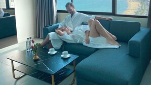 A modern living room with a large window offering a city view, a sofa, a coffee table, and a vase with flowers. - Hermann Hotel Pattaya (Pattaya)