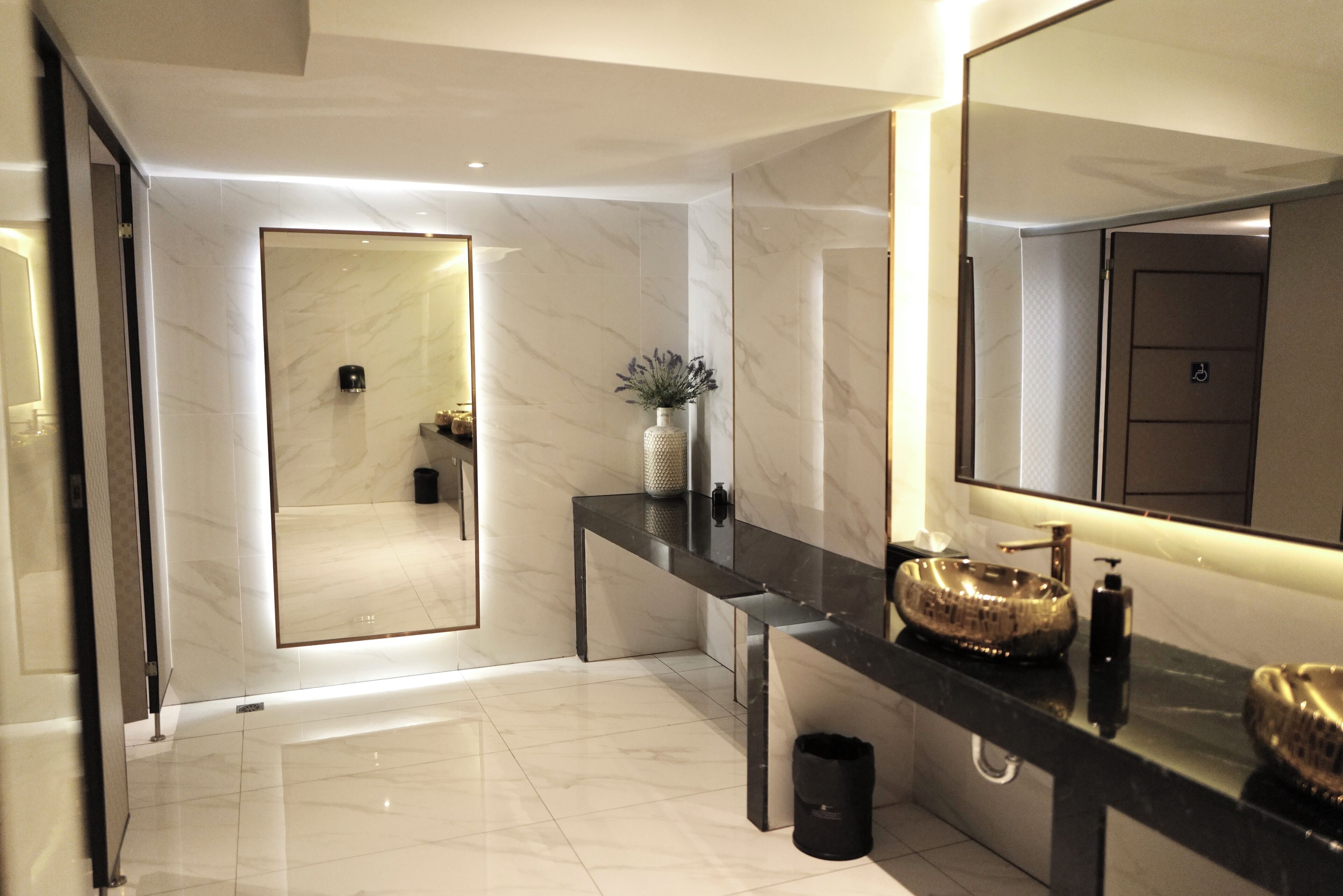 Un baño moderno con piso de mármol, un espejo grande, encimera negra y un cuenco decorativo dorado.
