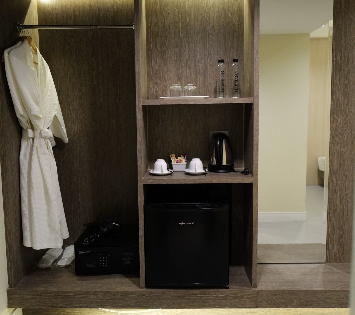 A hotel room with a wooden wardrobe, a small refrigerator, a kettle, and a robe hanging on the wall.