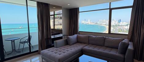 A modern living room with a large sectional sofa, a glass coffee table, and a view of the ocean and city skyline through large windows.