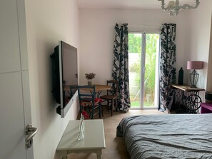 A hotel room with a bed, a TV mounted on the wall, a small table with a vase, a chair, a table with a lamp, and a sliding glass door leading to a balcony.
