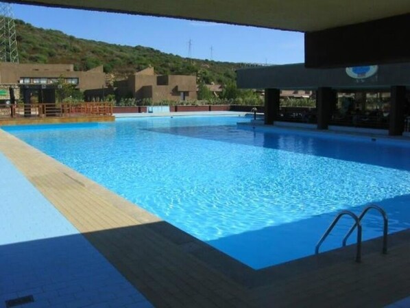A large swimming pool with a wooden deck area, surrounded by buildings with a hilly backdrop.