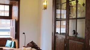 A cozy room with a wooden ceiling, a large cabinet with glass doors, a cushioned armchair, and a window with a view of buildings. - Emblematico San Agustin Hotel (Icod de los Vinos)