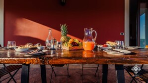 Una mesa de comedor de madera dispuesta con alimentos para el desayuno, incluyendo frutas, pasteles y jugo de naranja, frente a una pared roja con una puerta y una lámpara fijada a la pared.