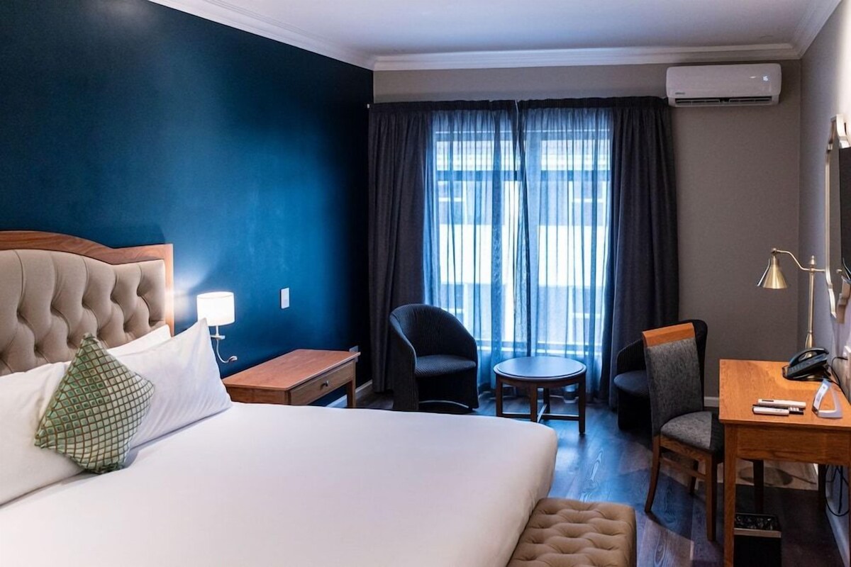 Une chambre d’hôtel comprenant un lit, un bureau, deux chaises, une petite table et une fenêtre avec des rideaux.