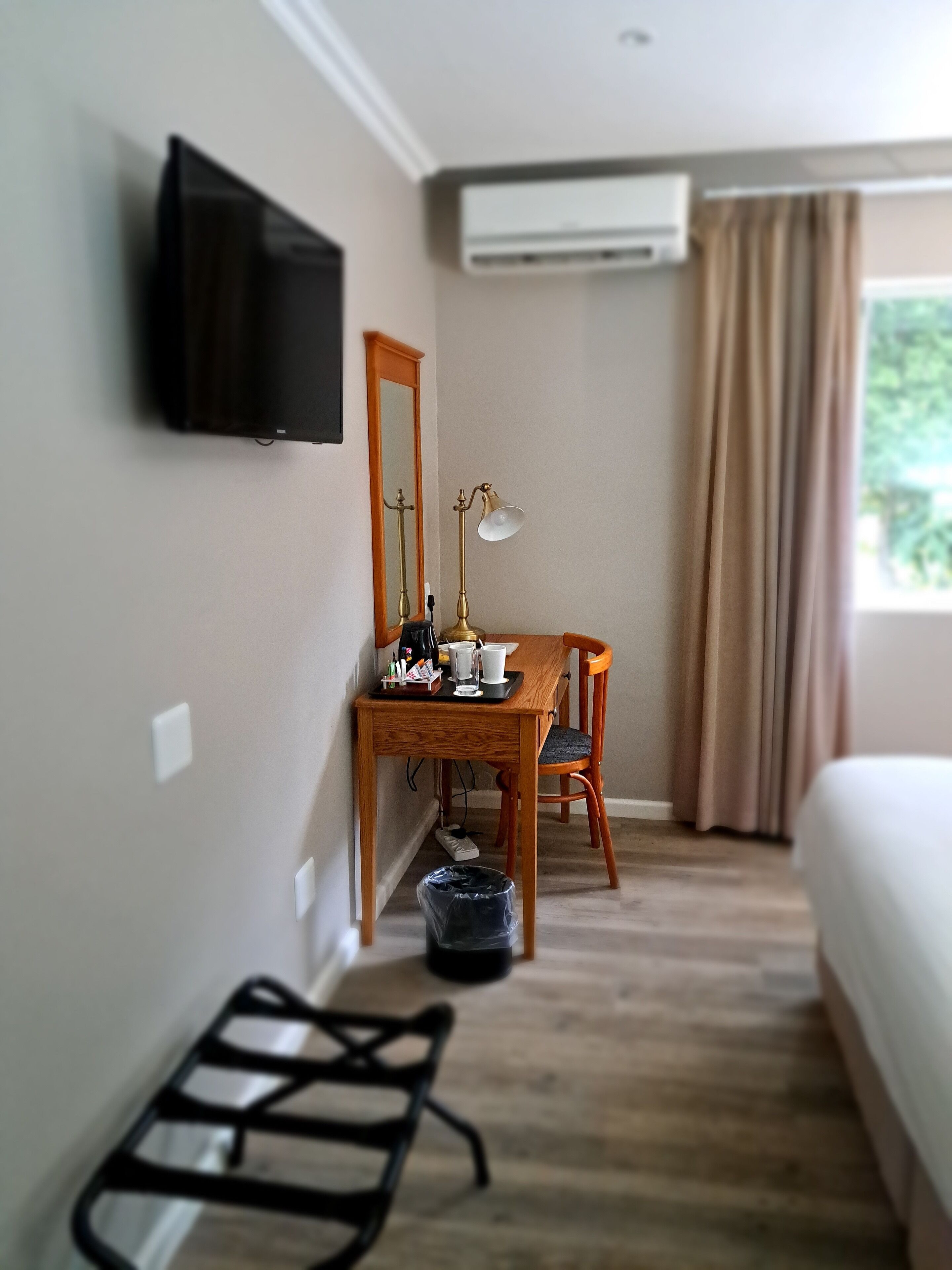 A hotel room with a wooden desk, a chair, a television mounted on the wall, an air conditioning unit, and a window with curtains.