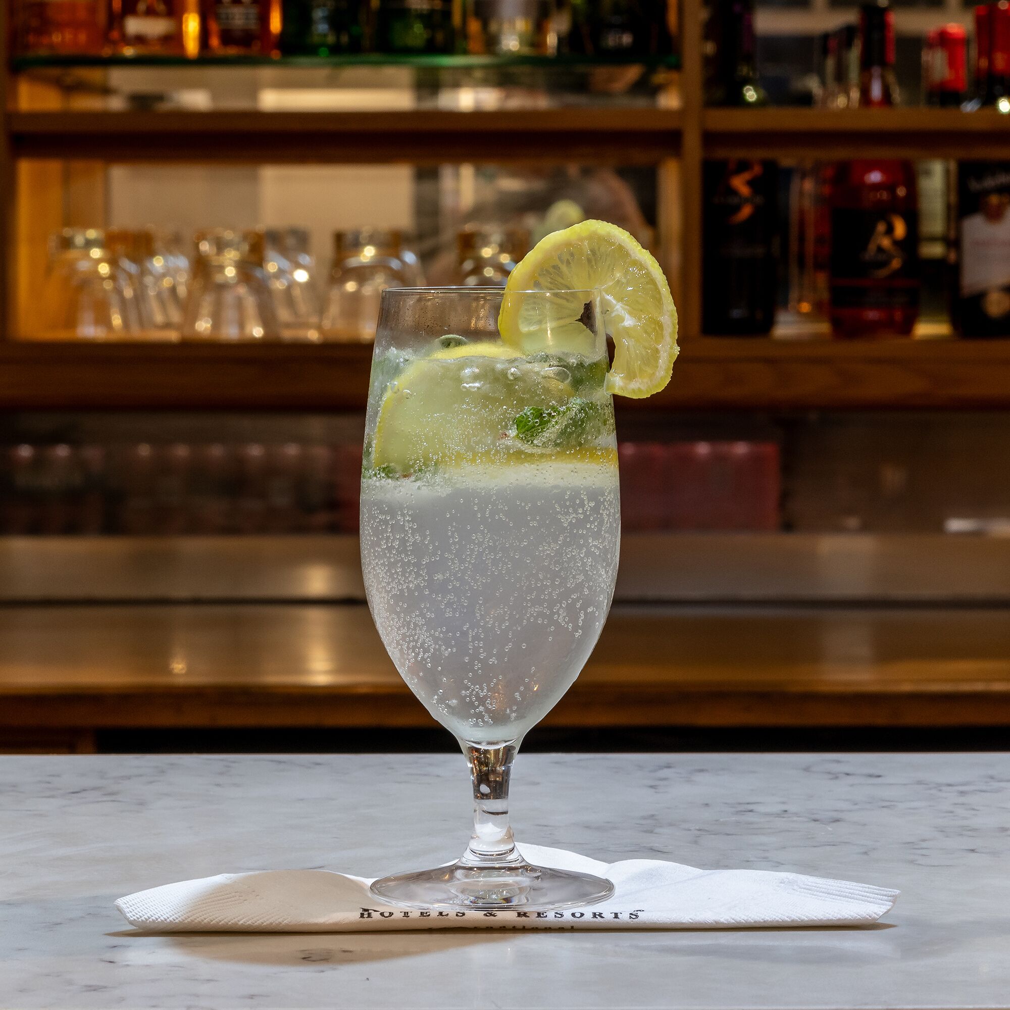 A bar setting with a glass of a clear, bubbly beverage garnished with a slice of lemon, placed on a marble surface with a napkin underneath.